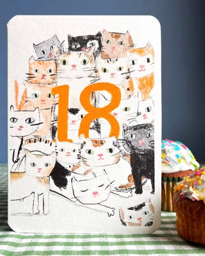 Greeting card with cat illustrations and the number 18 on a table with cupcakes.