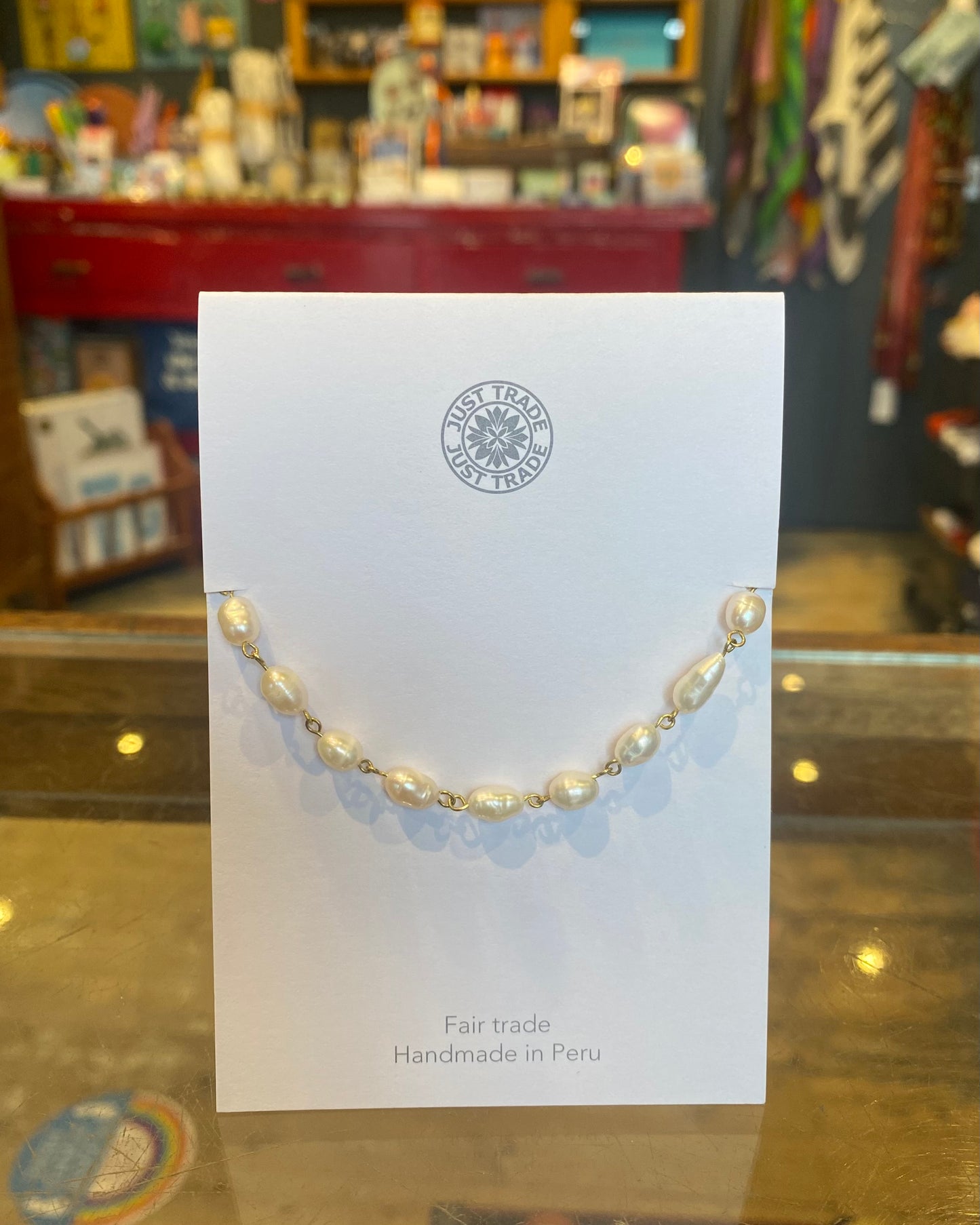 Pearl bracelet on a white card with 'Fair Trade' and 'Handmade in Peru' text, displayed against a blurred store background.