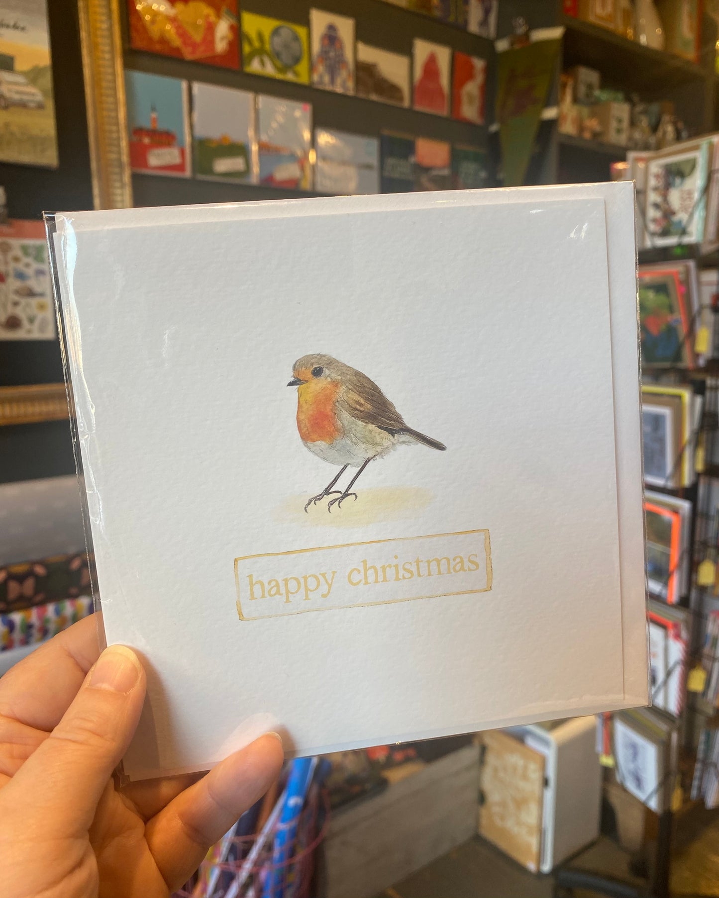 Card with a robin illustration and 'happy christmas' text held in front of a bookshelf.