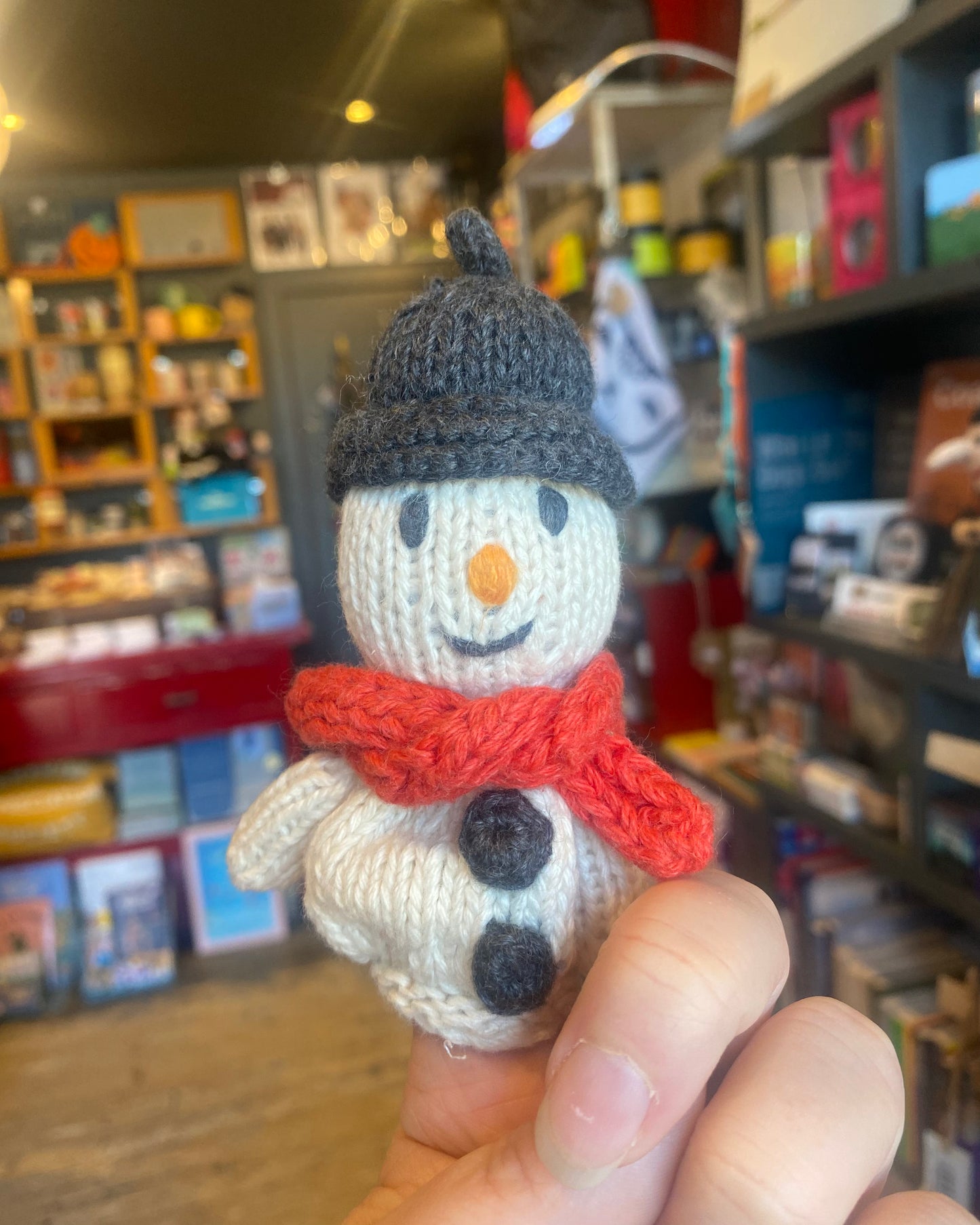 Knitted snowman finger puppet on a hand, with a store interior in the background.