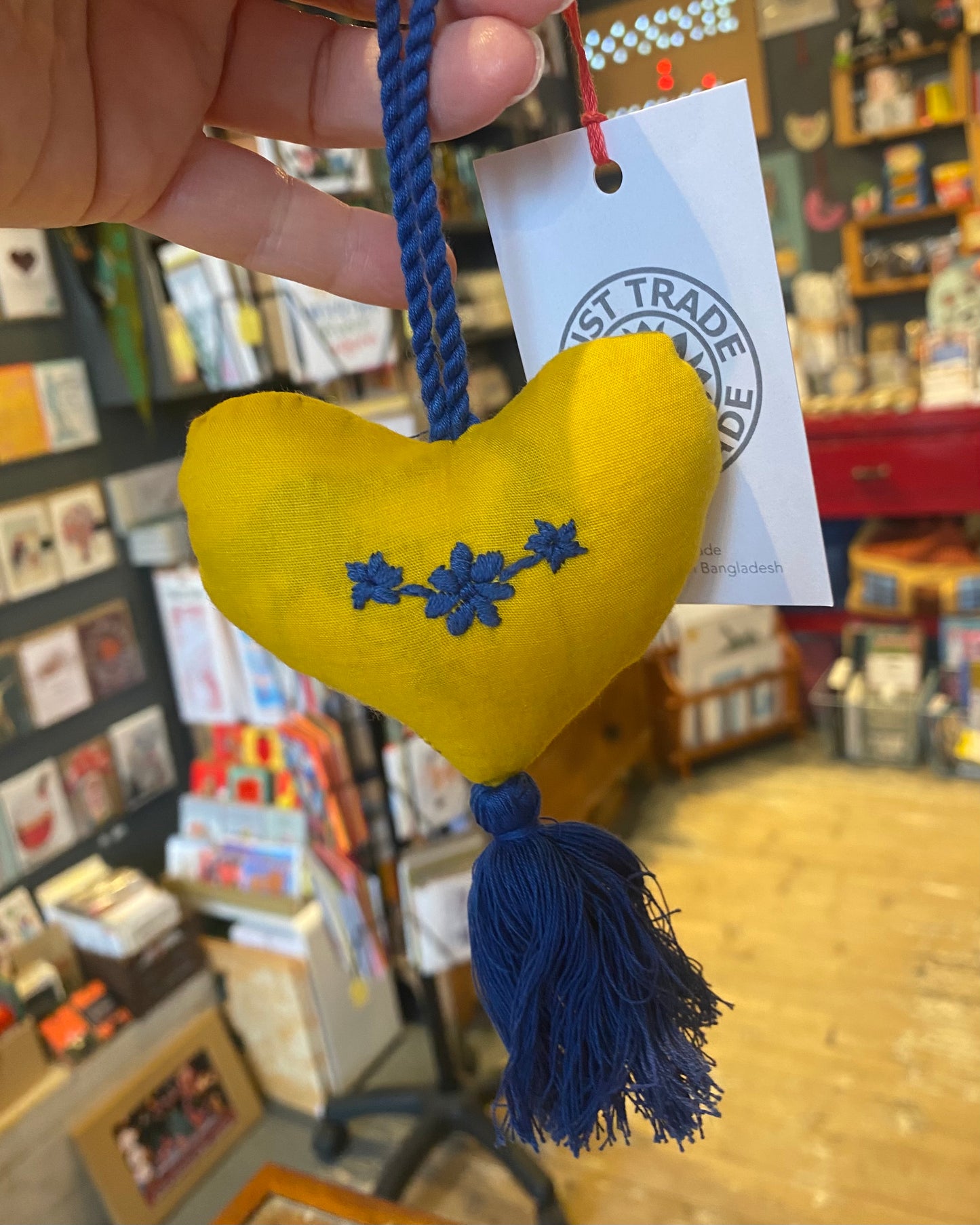 Yellow heart-shaped plush decoration with blue tassel and embroidered design, held by a hand with a visible 'Just Trade' label.