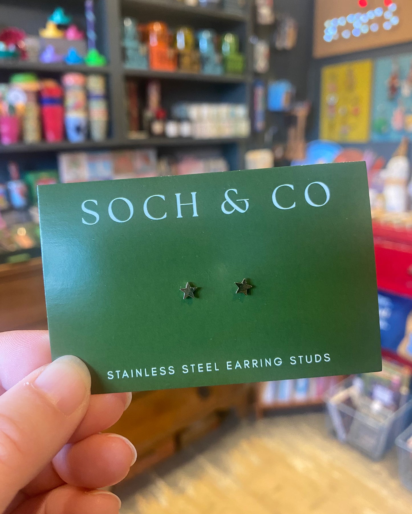 Star-shaped earrings on a green card with 'SOCH & CO' branding, held in front of a store shelf.