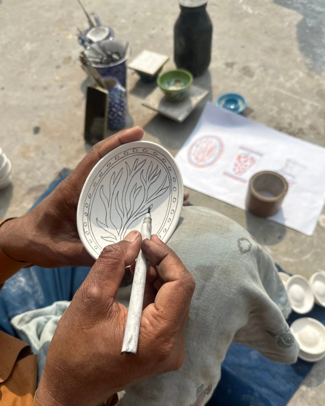 Person holding a ceramic soap dish with drawn design and a small tool, with a workshop setting in the background.