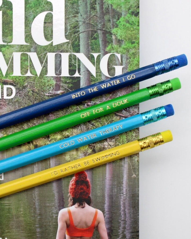 Four pencils with motivational phrases about wild swimming, on a magazine cover featuring a forest and swimmer.