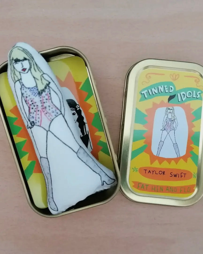 Open metallic tin with illustration of Taylor Swift and text 'Tinned Idols Taylor Swift', with sewn mini Taylor inside, on a wooden surface.