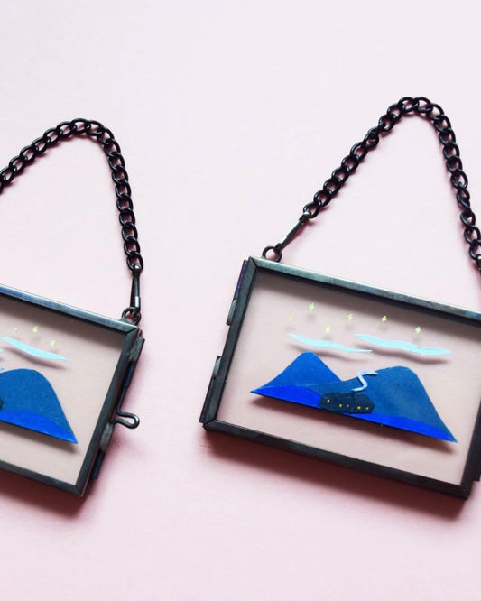 Two framed mini glass frames with blue mountain and both designs on a pink background