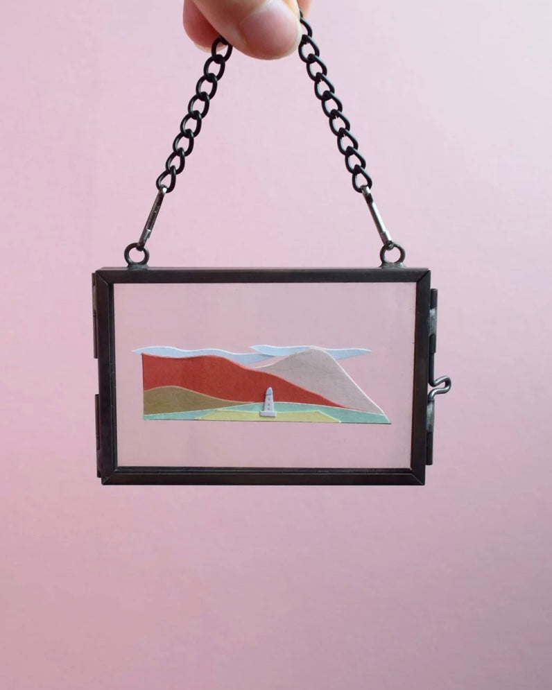 Small framed artwork with a mountain and lighthouse scene on a pink background