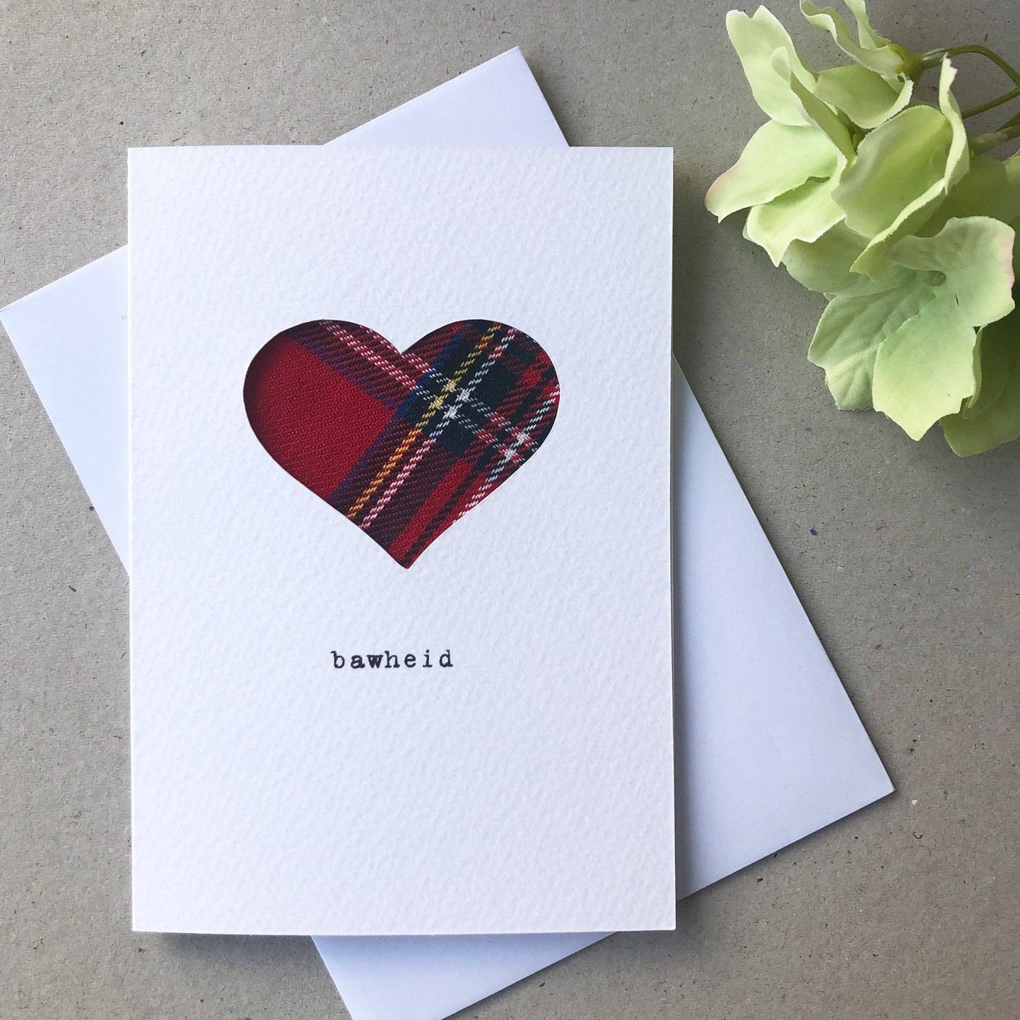 White card with a real cloth tartan heart design and 'bawheid' text, placed on a white envelope, on a textured surface with a flower.