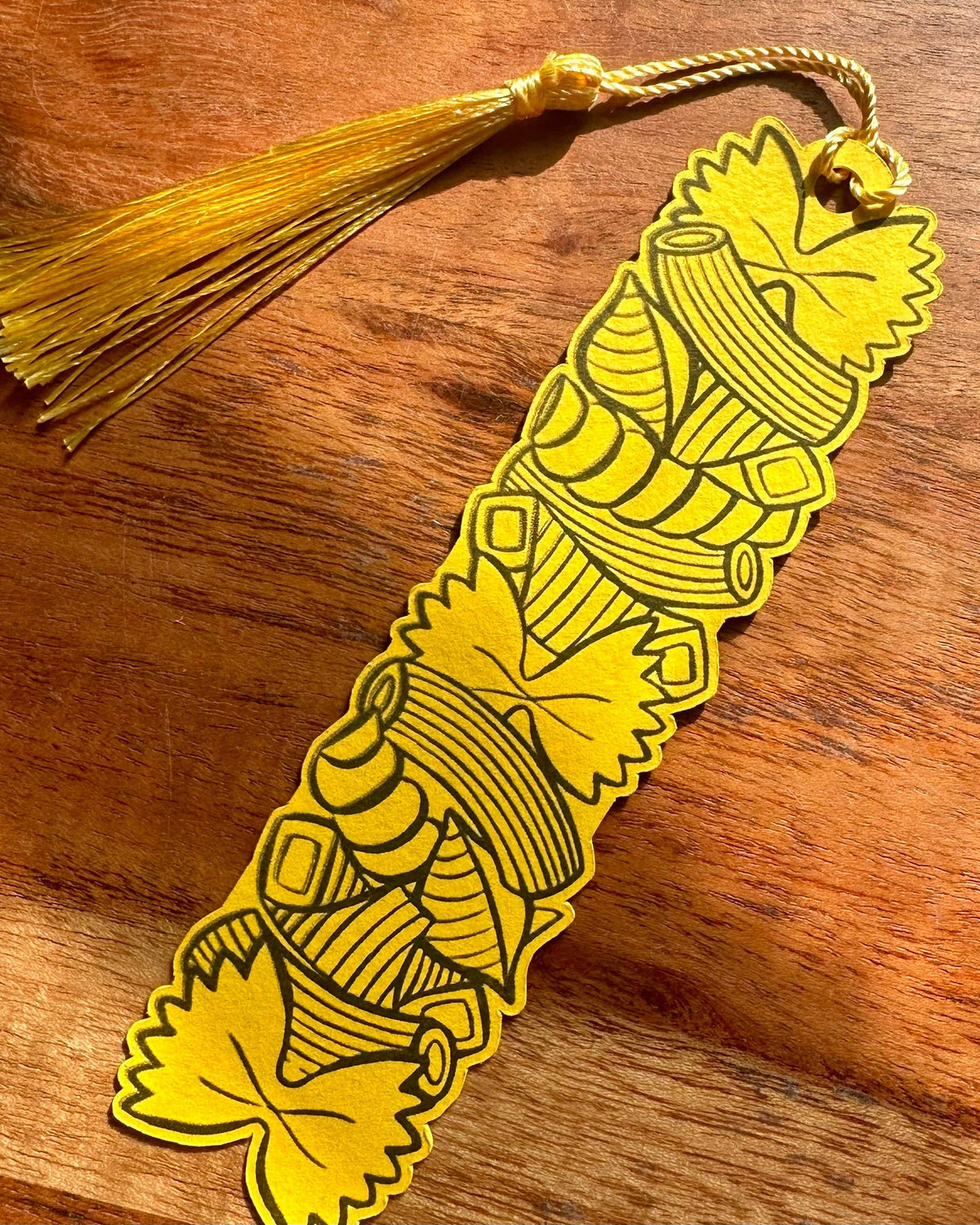 A yellow paper bookmark with black pasta shape patterns and a matching yellow tassel, resting on a wooden surface.