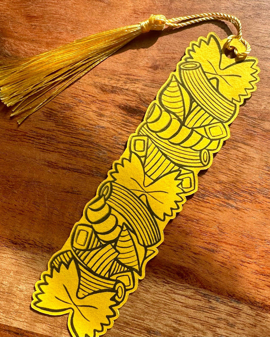 A yellow paper bookmark with black pasta shape patterns and a matching yellow tassel, resting on a wooden surface.