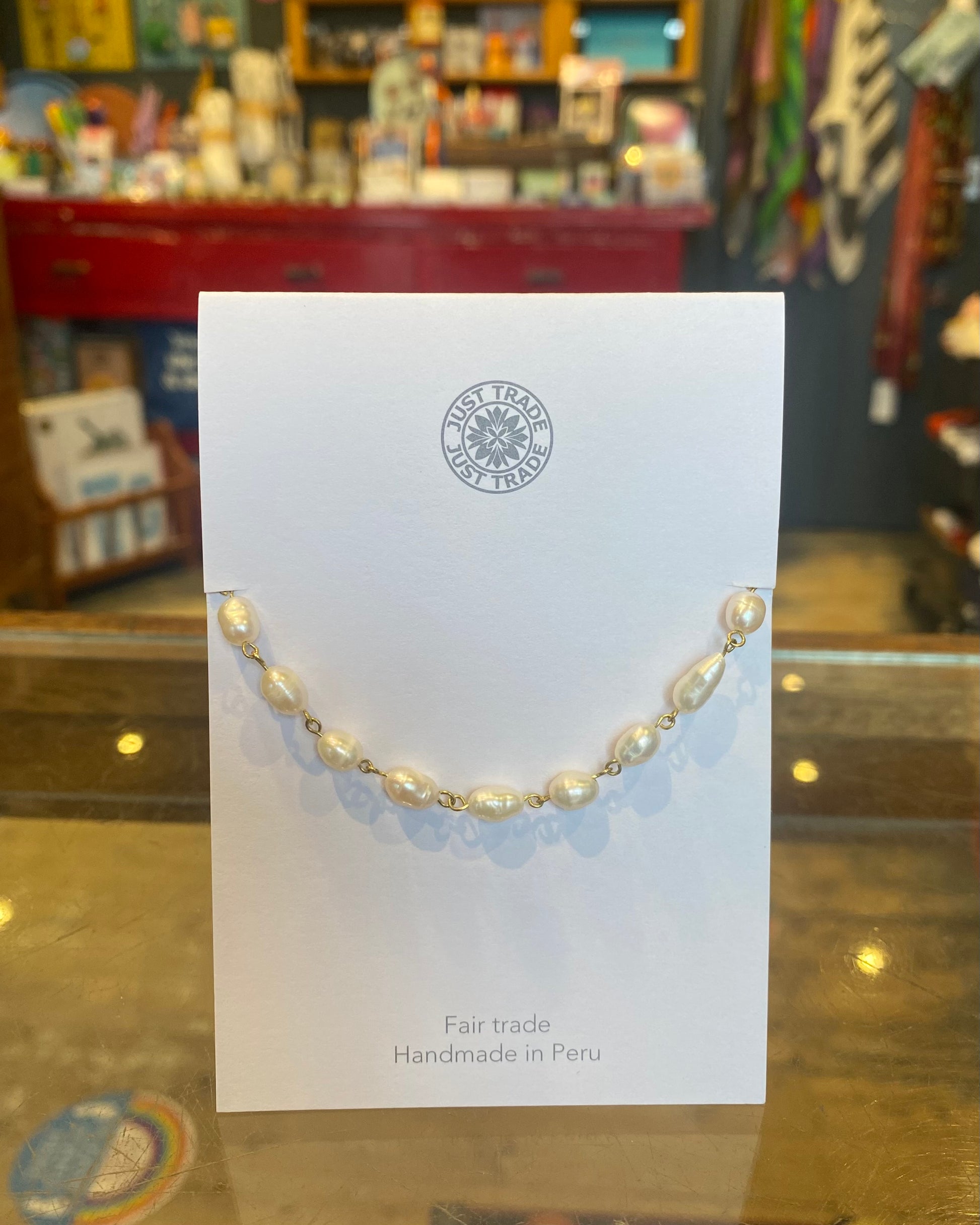 Pearl bracelet on a white card with 'Fair Trade' and 'Handmade in Peru' text, displayed against a blurred store background.