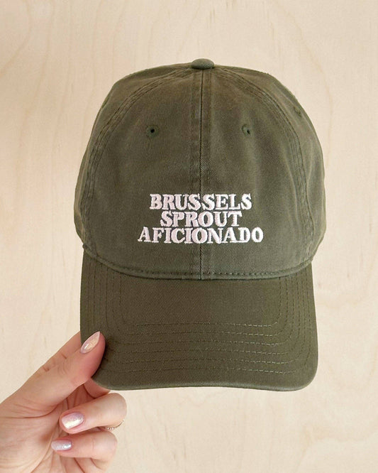 Green cap with 'Brussels Sprout Aficionado' text held by a hand on a beige background