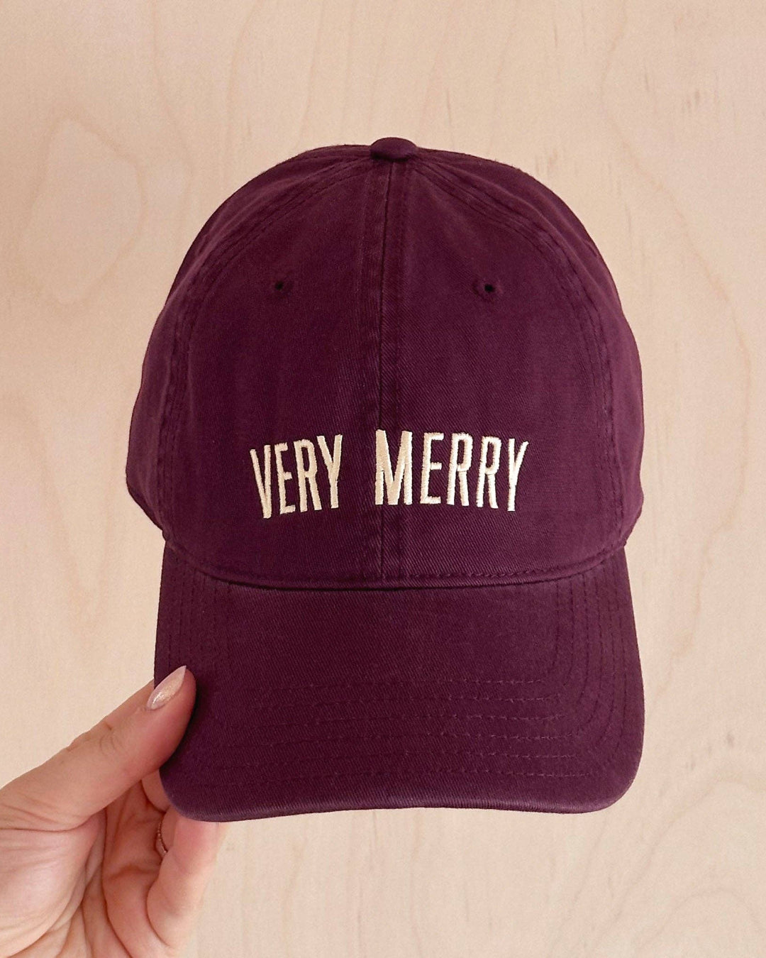 Maroon cap with 'VERY MERRY' text held by a hand on a beige background