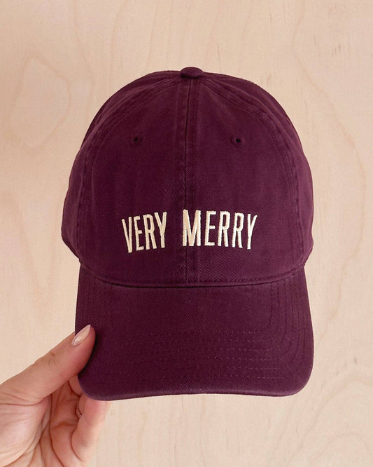 Maroon cap with 'VERY MERRY' text held by a hand on a beige background