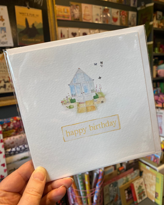 Hand holding a birthday card with a greenhouse illustration in a store setting