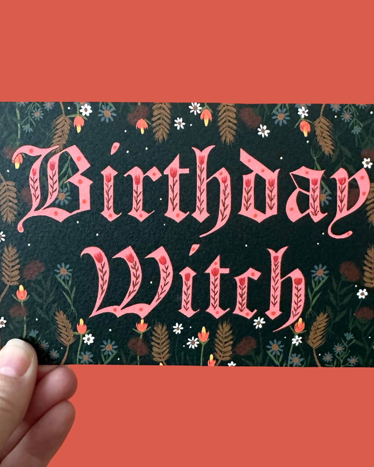 A hand holds a greeting card with the words “Birthday Witch” in pink decorative lettering over a dark forest-patterned background with small flowers and leaves, against a solid coral-red backdrop.