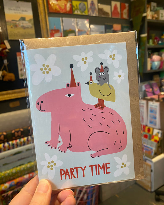 Card with a pink capybara, bird & mouse illustration and 'Party Time' text, held in front of a store shelf.