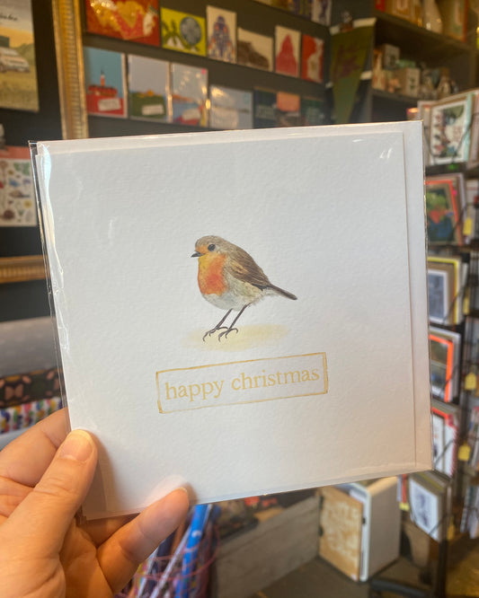 Card with a robin illustration and 'happy christmas' text held in front of a bookshelf.
