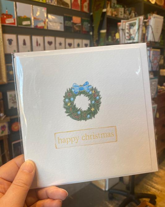 White card with Christmas wreath design held in a hand, with a store interior in the background.