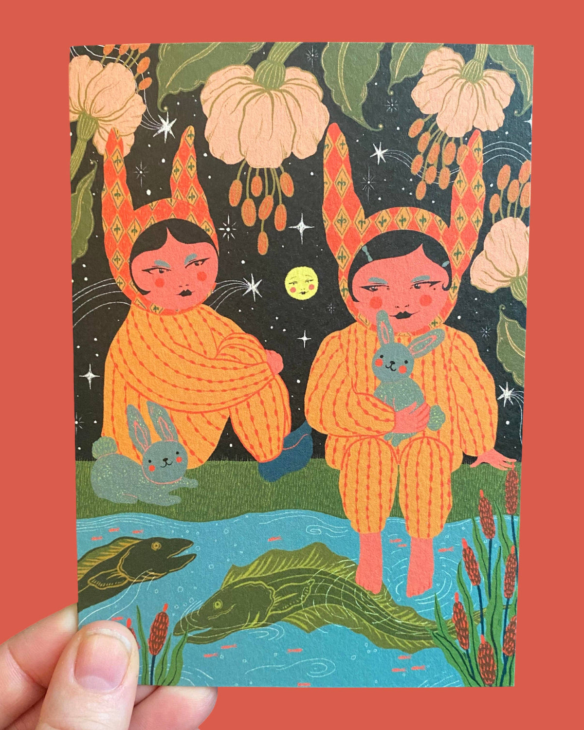 A hand holds a card featuring a whimsical illustration of two children in orange bunny suits sitting by a pond under a starry night sky with pumpkins and flowers. The card is set against a solid coral background.