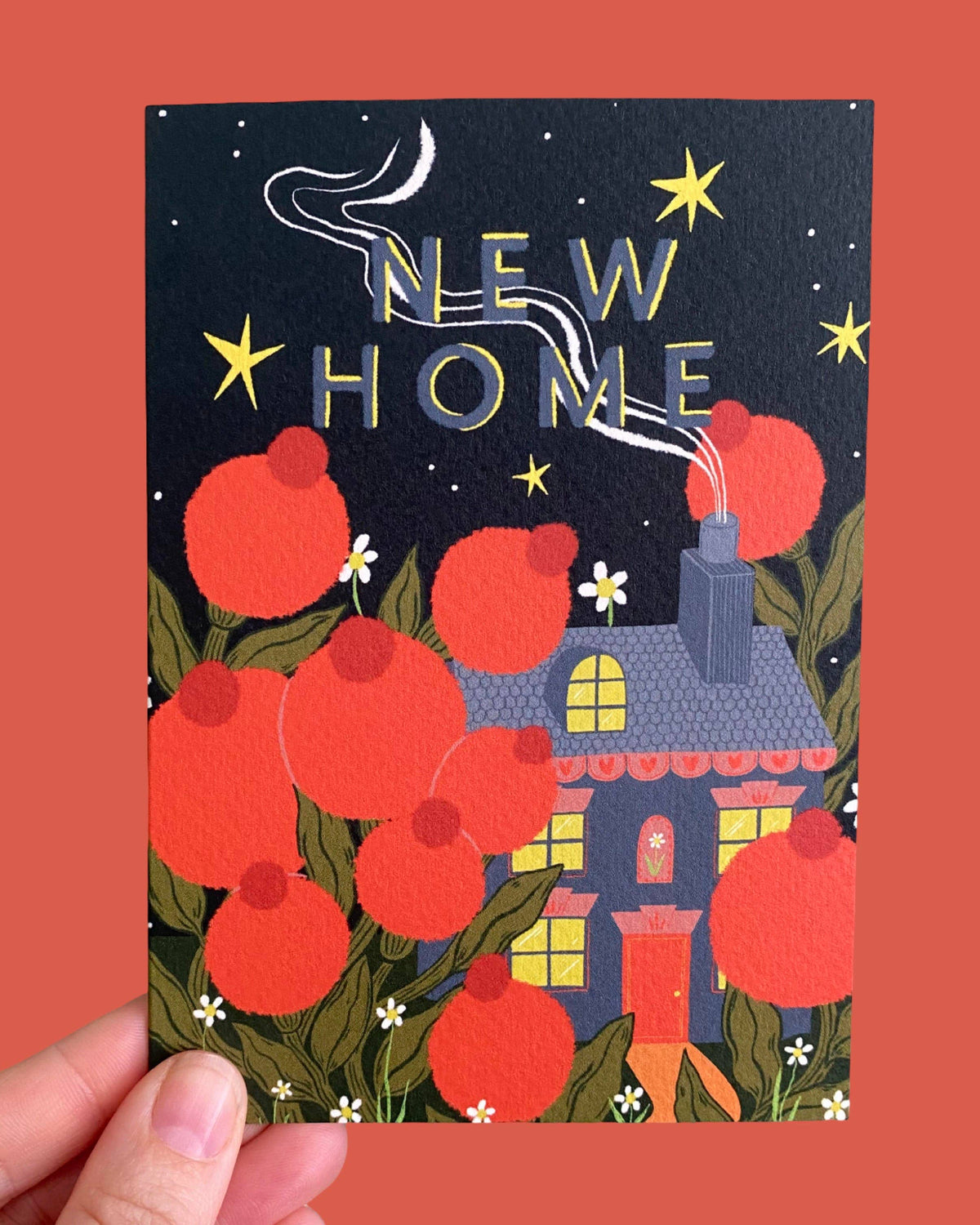 A hand holds a greeting card featuring a whimsical illustration of a house, large red flowers, green leaves, and the words 'NEW HOME' in gold foil against a dark night sky. The card is shown against a solid coral-red background.