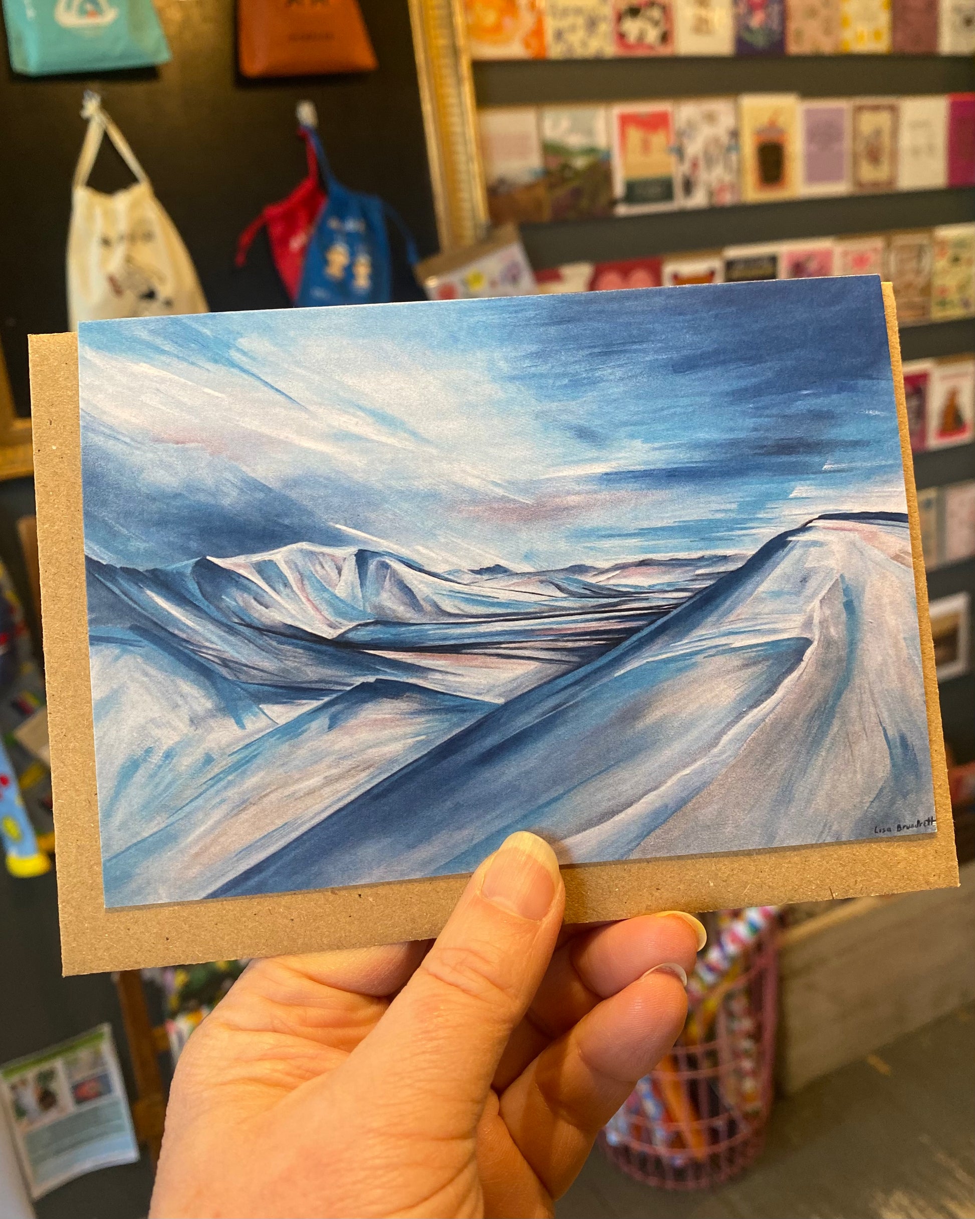 Hand holding a small painting of a snowy landscape with blue skies and white clouds.