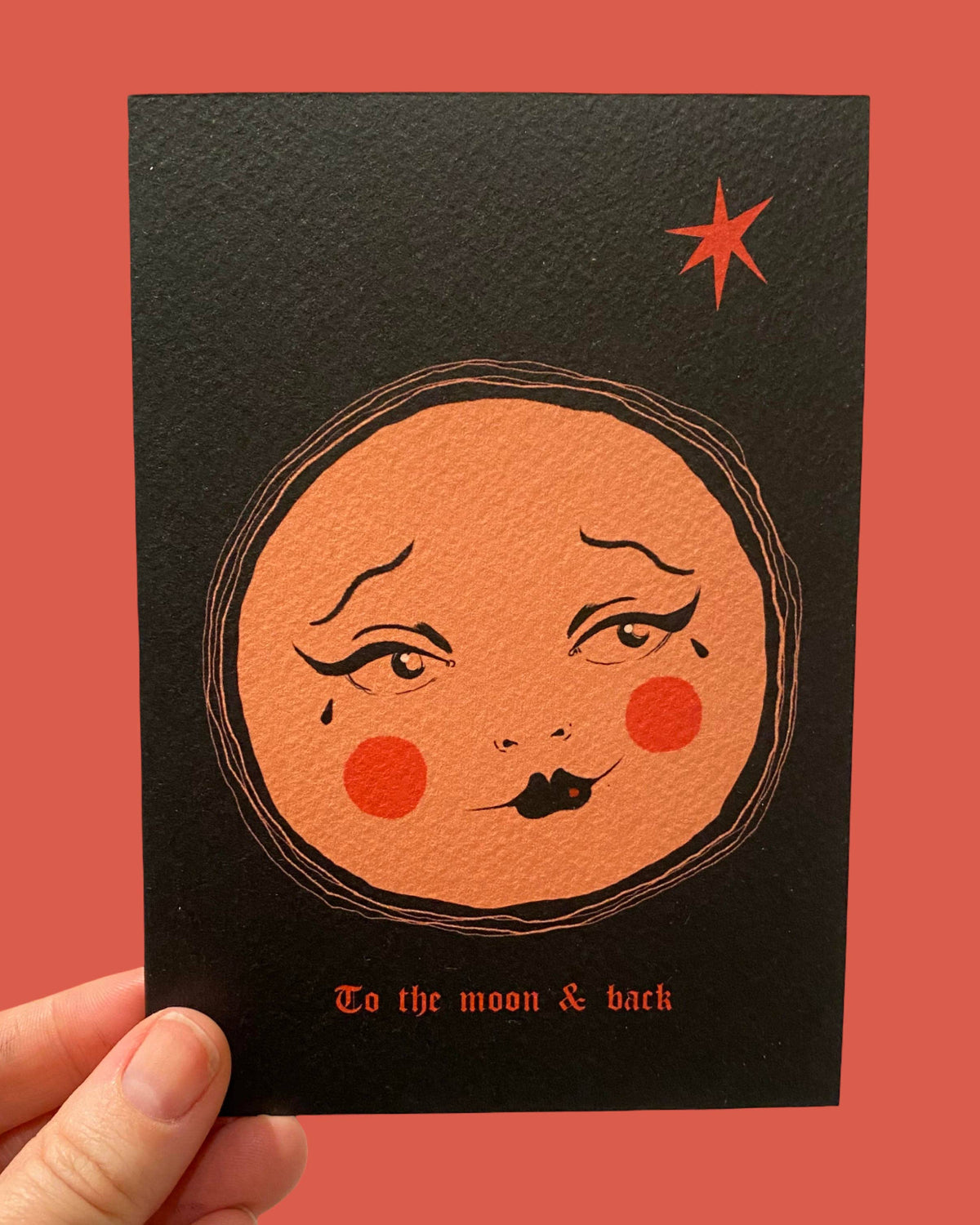 A hand holds a black art print card featuring a stylized orange moon face with red cheeks and the text 'To the moon & back' against a solid coral background.