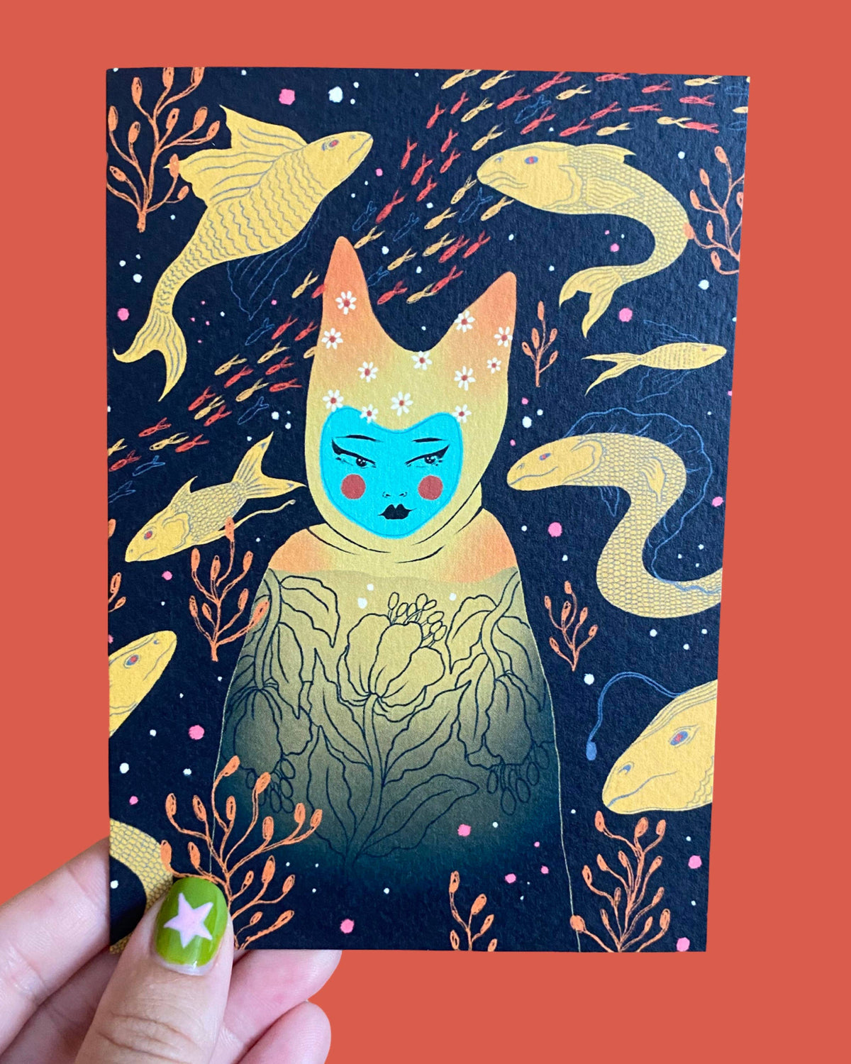 Card with a cat-like creature illustration on a dark blue background with gold fish and plants, held against an orange background.