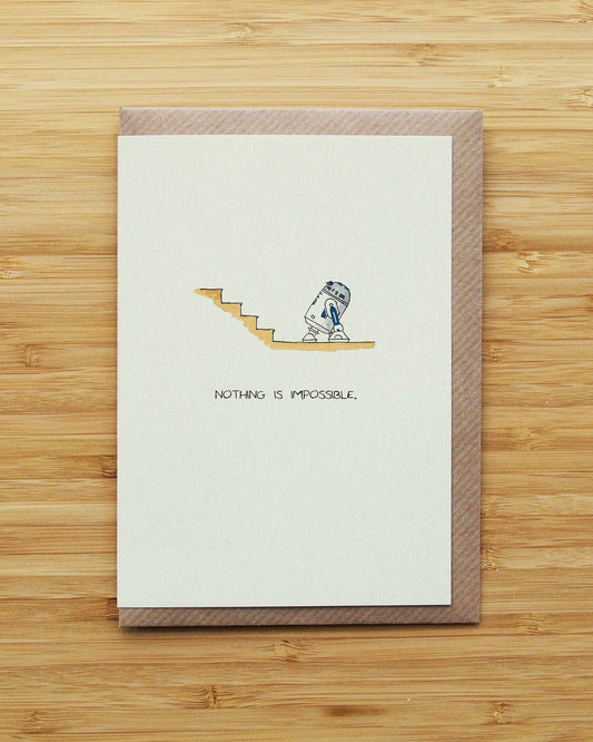 A  greeting card with a brown envelope, featuring a small illustration of a blue-and-white robot (R2D2) climbing a tan staircase and the text 'NOTHING IS IMPOSSIBLE.' on a light wood background.