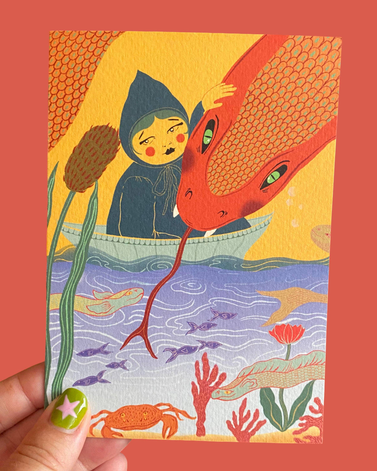 A hand holds a square art print featuring a whimsical illustration of a child in a boat with a large red serpent, set against a coral-red background.