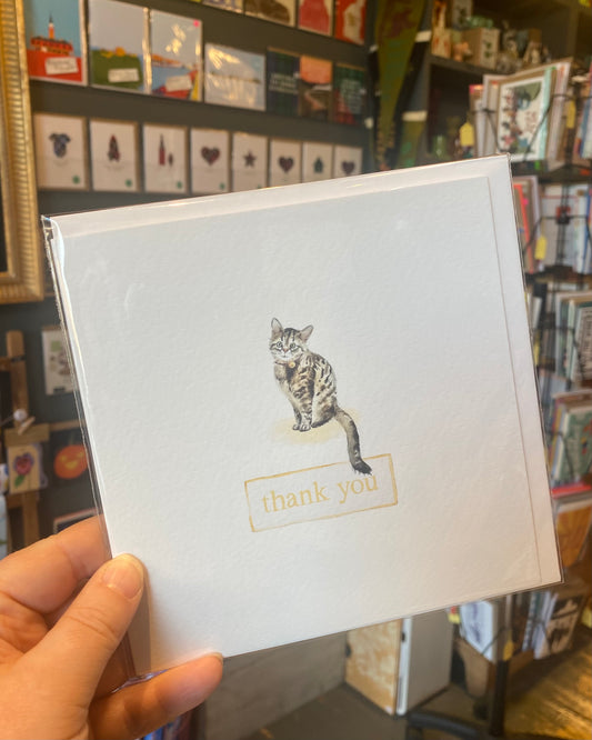 Card with a cat illustration and 'thank you' text held in front of a bookshelf.