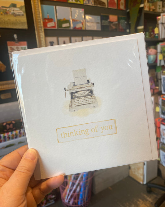 White card with typewriter illustration and 'thinking of you' text held in a hand with a store background.