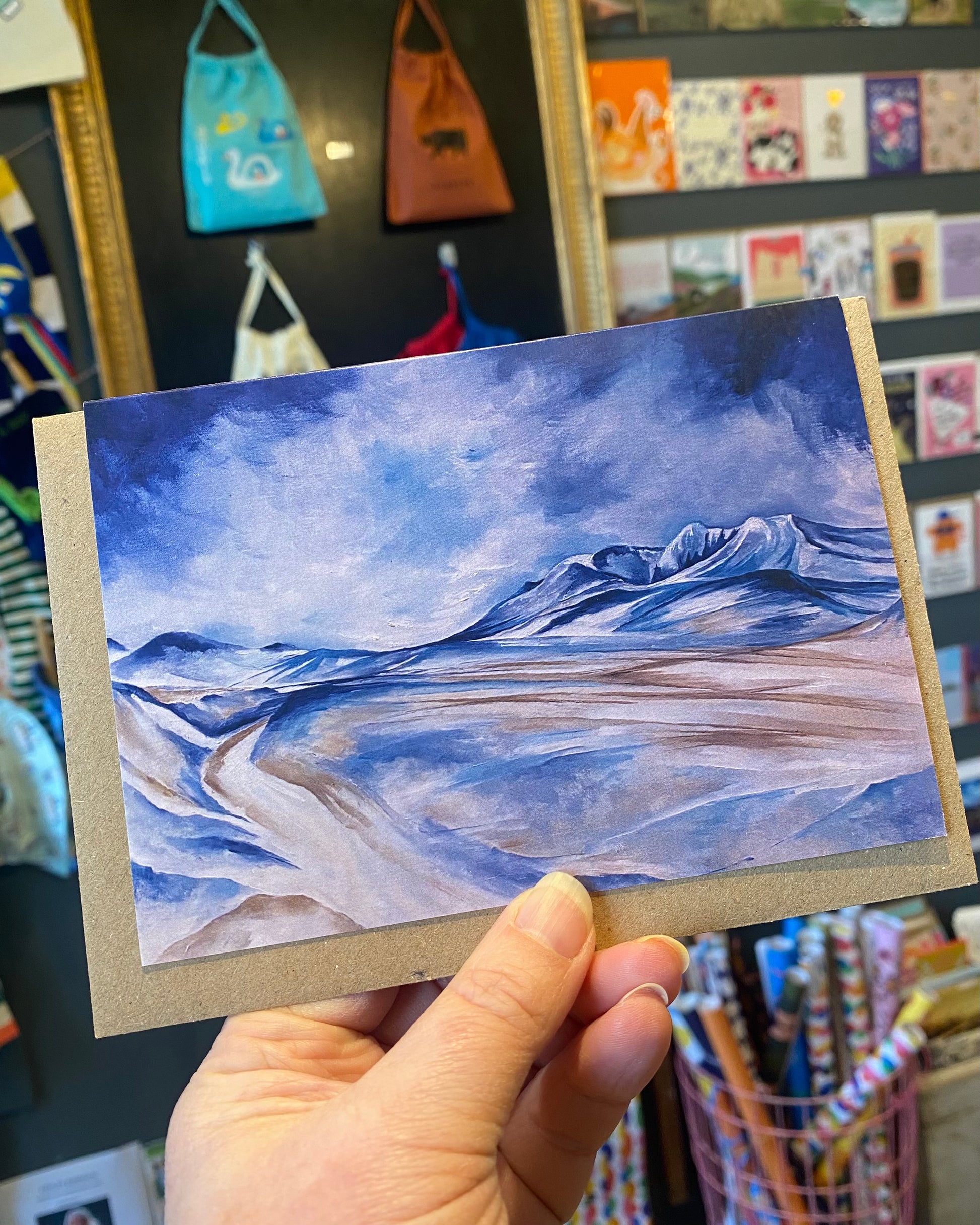 Hand holding a small landscape painting with blue and brown tones.