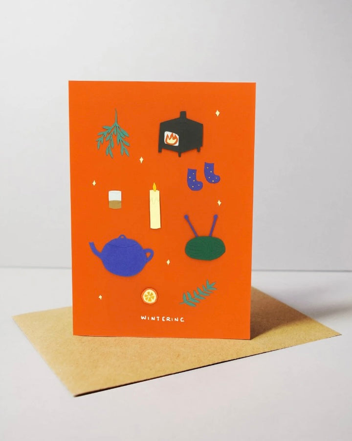 Greeting card with illustrations on an orange background, featuring a teapot, boots, and other winters objects, with a brown envelope below.