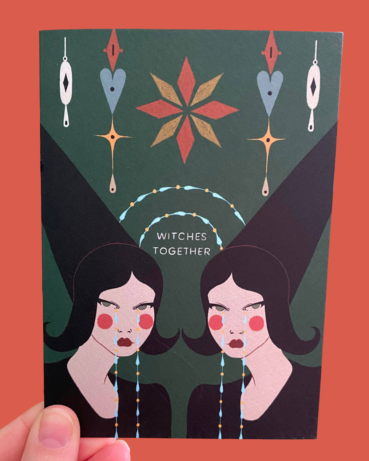 A hand holds a square art card featuring a stylised illustration of two women with pointed hats and the text 'WITCHES TOGETHER' on a dark green background, set against a solid coral-red backdrop.