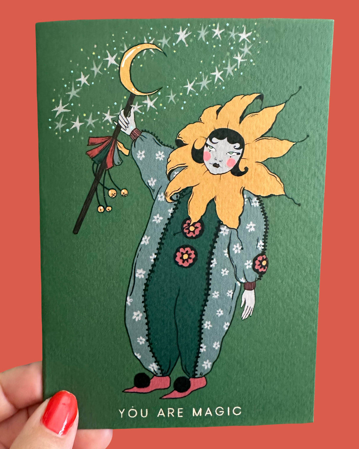 A hand holds a green illustrated card featuring a whimsical sunflower-like character with a crescent moon and stars above, with the text 'YOU ARE MAGIC' at the bottom, against a coral-red background.