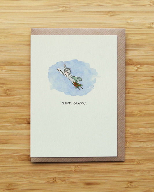 A greeting card with a brown textured border and a small watercolour illustration of a woman flying like a superhero under the text 'SUPER GRANNY', displayed on a light wood background.