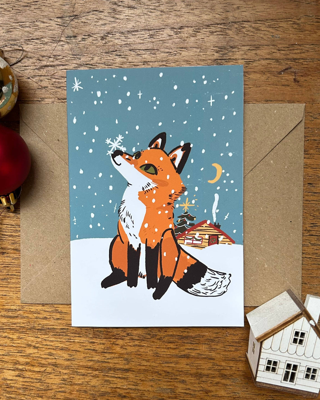 Greeting card with a fox illustration on a snowy night, surrounded by Christmas decorations on a wooden surface.
