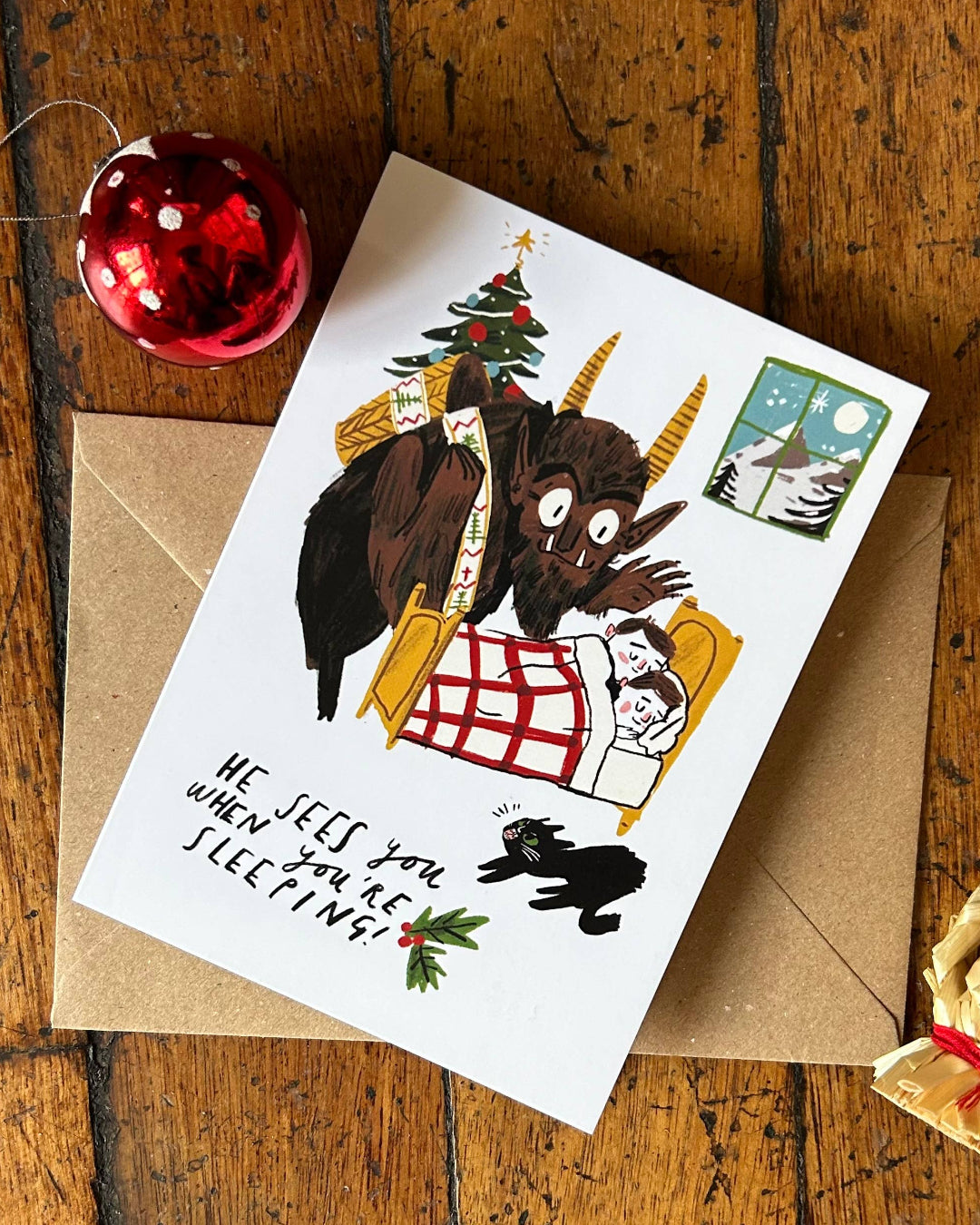 Christmas card with cartoon Krampus design and 'He sees you when you're sleeping' text on a wooden surface with festive decorations.