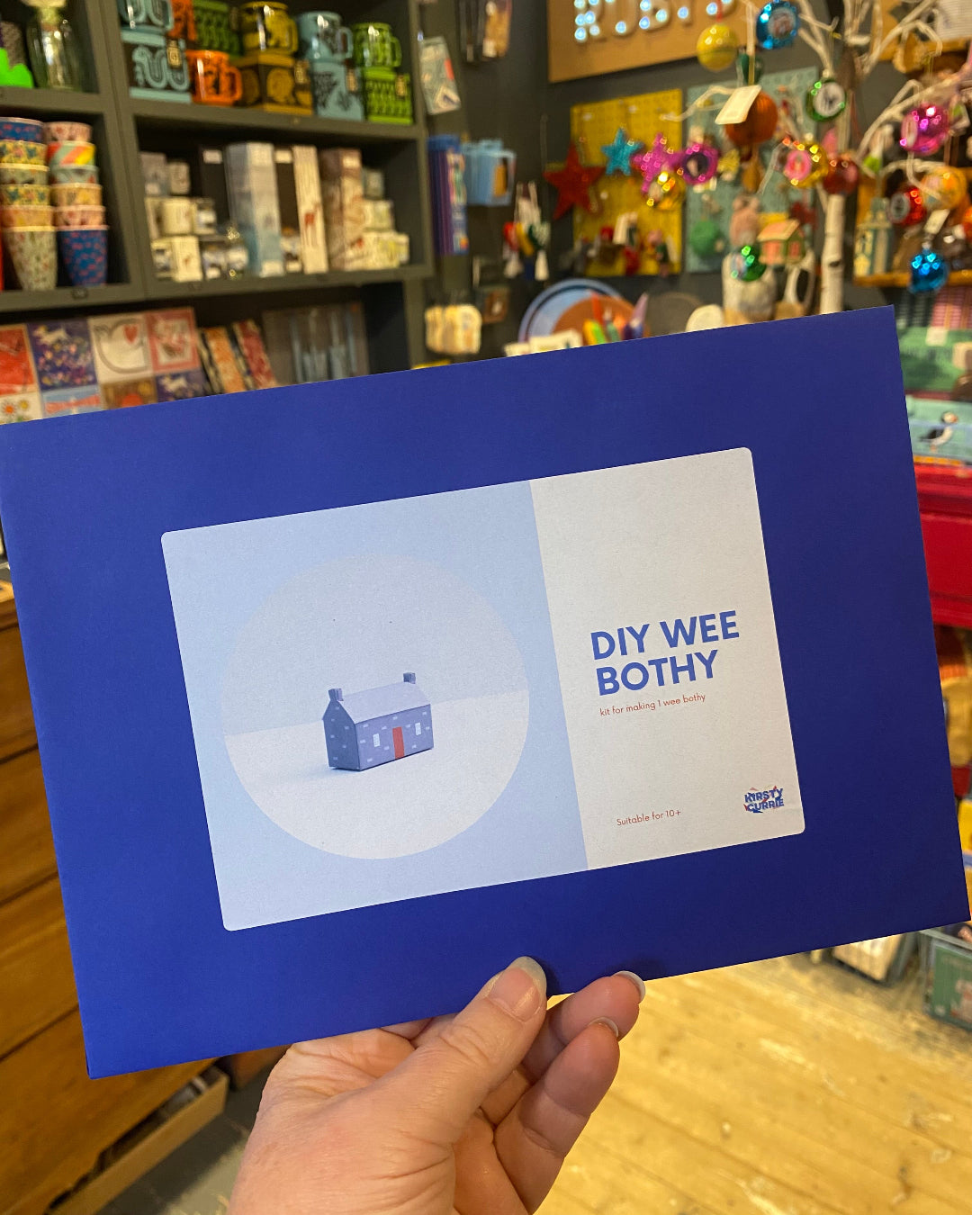 Blue envelope with a small house (both) DIY kit held in front of a store shelf.