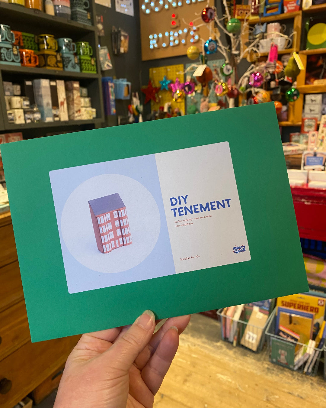 Hand holding a green card with 'DIY Tenement' design in a store setting