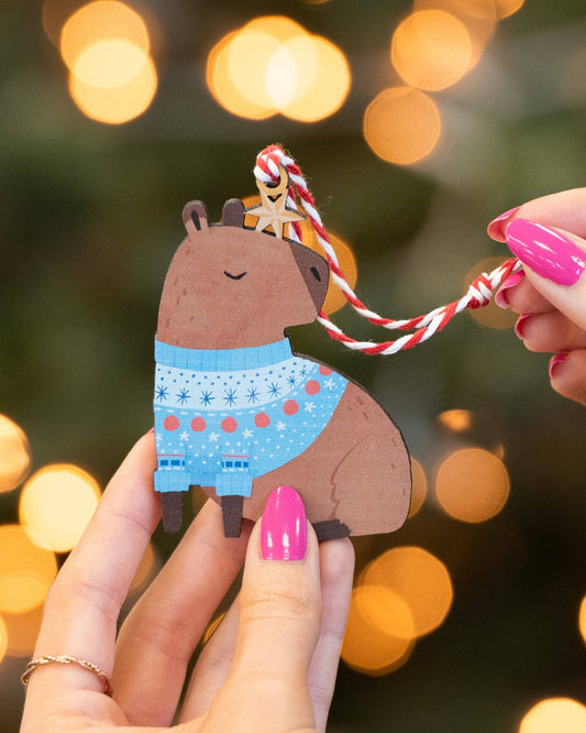 A small wooden capybara ornament with a blue patterned sweater and a red-and-white hanging string, held in two hands against a blurred festive background with warm bokeh lights.