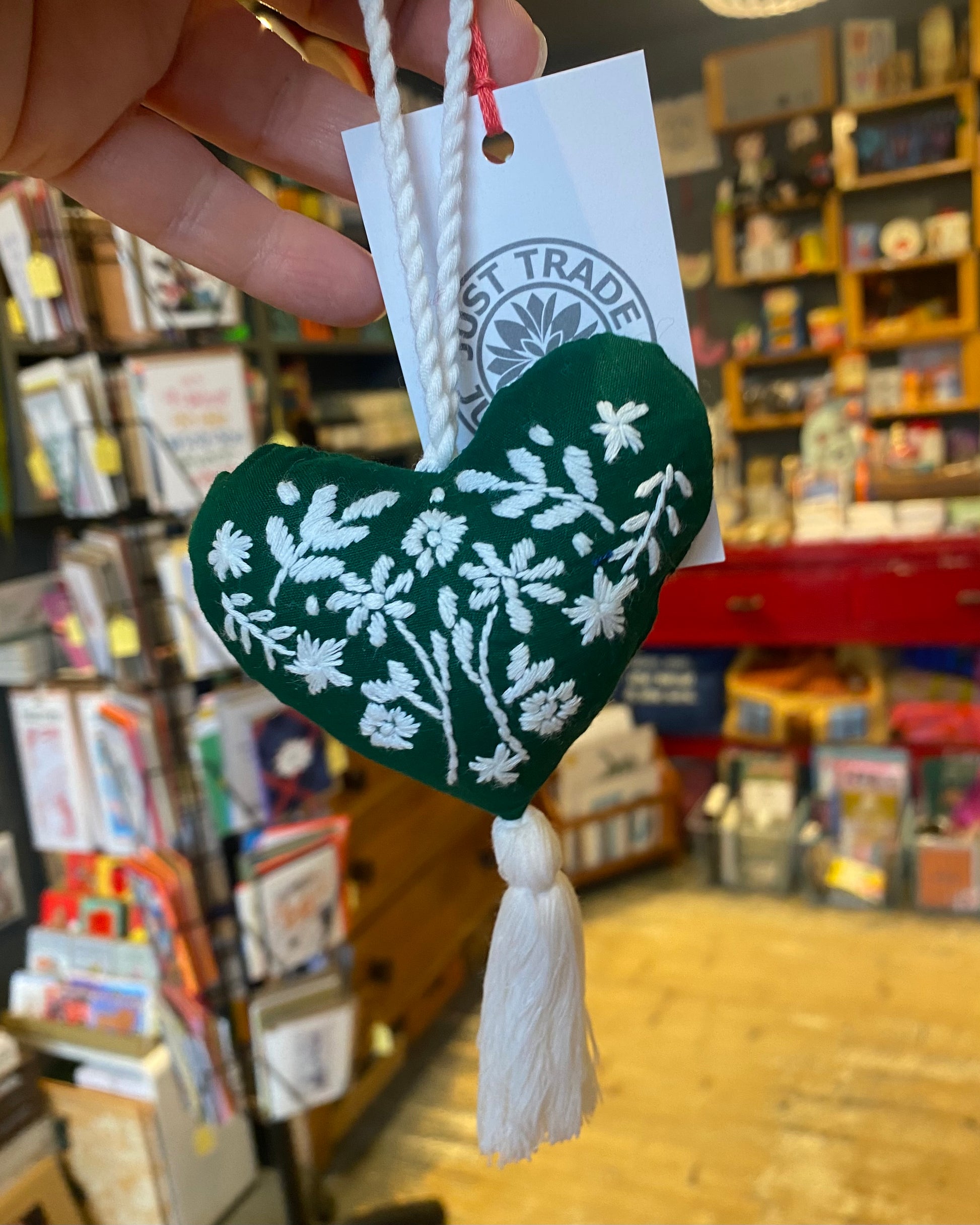 Green heart-shaped ornament with white floral patterns on a tag, held in a store setting.
