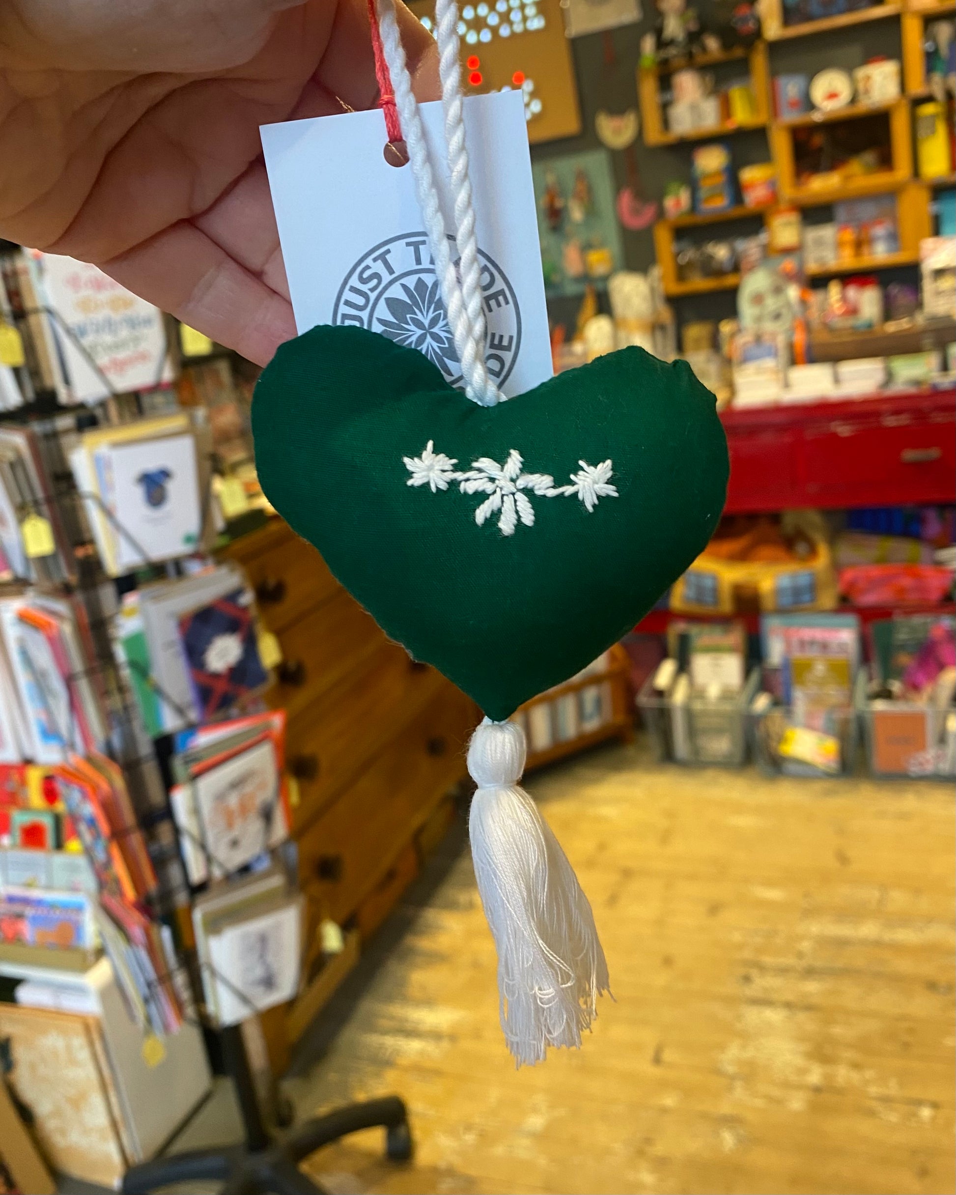 Heart-shaped green ornament with white floral design and tassel, held in front of a store display.