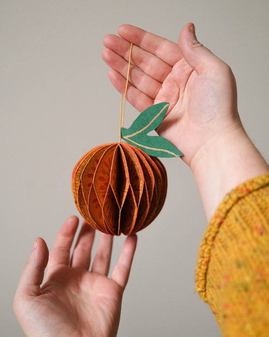 Hand holding a decorative paper fruit ornament against a neutral background