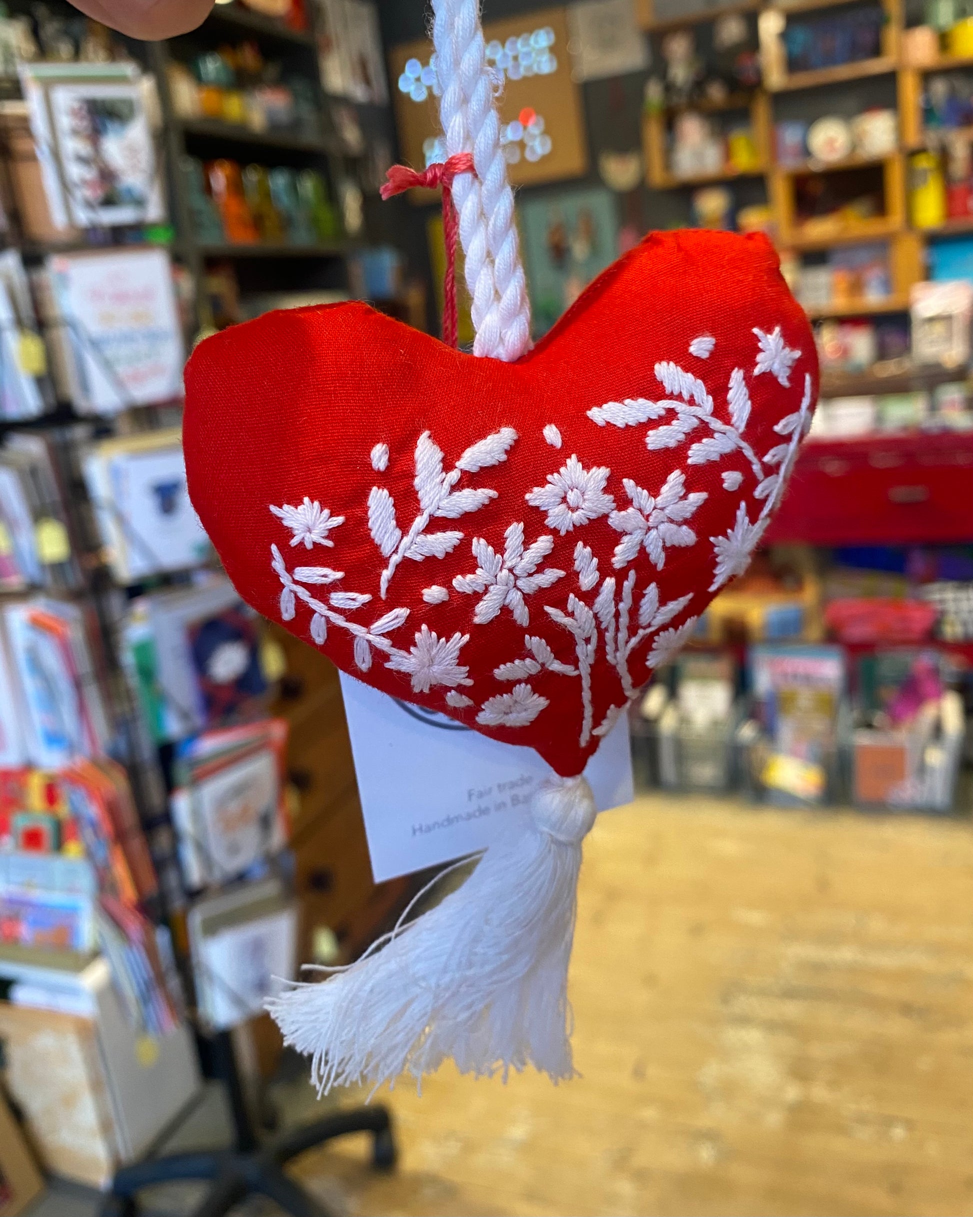Red heart-shaped textile ornament with white embroidery and tassle in a store setting