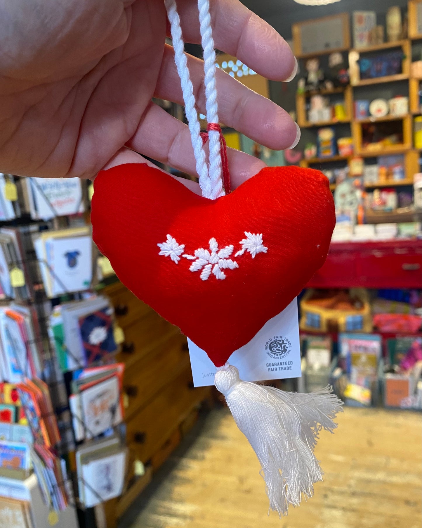 Red heart-shaped ornament with white embroidery and tassel, held in a hand with a store in the background.