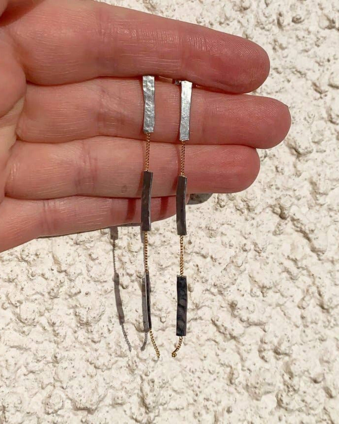 Hand holding a pair of long metal earrings against a textured beige background