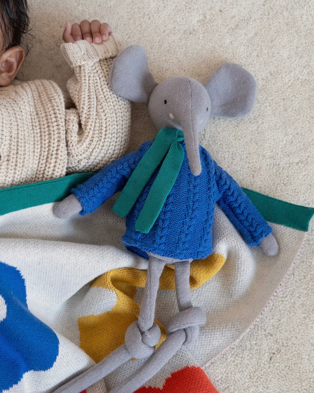 Toy elephant in a blue sweater on a colourful blanket with a child's hand in the background.