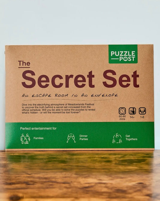 The Secret Set escape room puzzle envelope box by Puzzle Post, featuring a brown kraft paper design with green accents and text, displayed on a wooden surface against a light grey background.